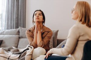 woman patient sits on couch appearing contemplative about addiction treatment programs while therapist sits near her discussing the patients options