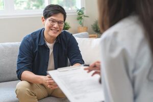 smiling person with glasses sits on a couch while listening to medical professional read of her clipboard in the persons depression treatment